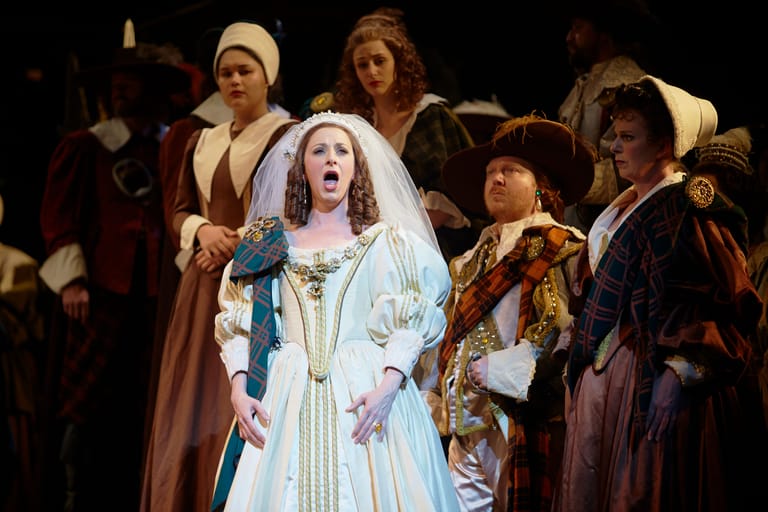 Female opera singer in pale blue wedding gown centre stage surrounded by costumed ensemble cast.