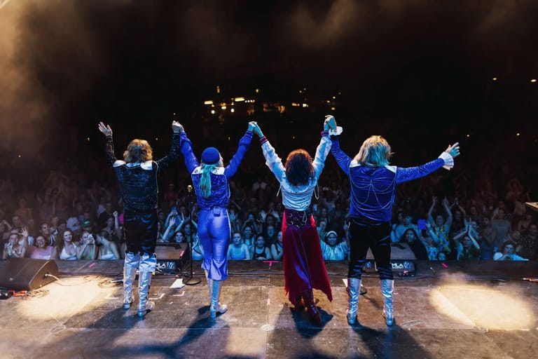 Five performers in blue clothing stand with arms raised in celebration on a lit stage before a crowd.