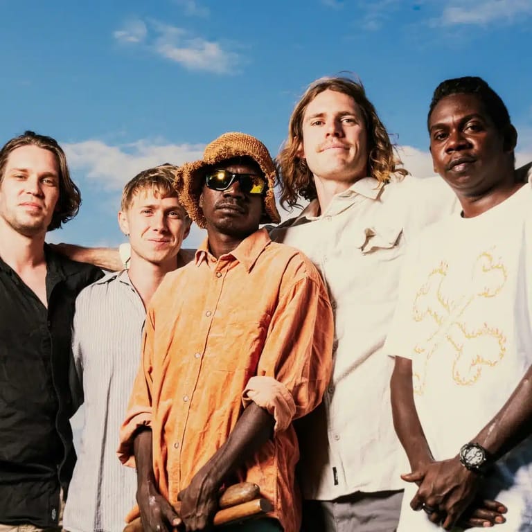 Five musicians stand together against a blue sky, wearing casual earth-toned clothing and sunglasses.