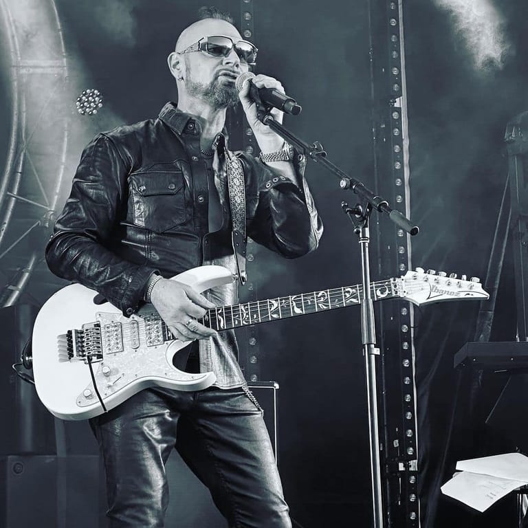 Performer wearing glasses and black leather jacket singing into microphone while playing white electric guitar on stage.