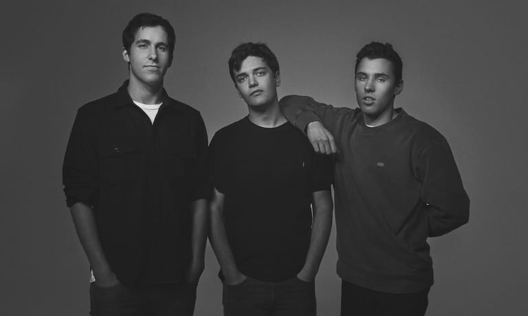 Three men in black shirts stand shoulder-to-shoulder against a grey background in a black and white portrait.