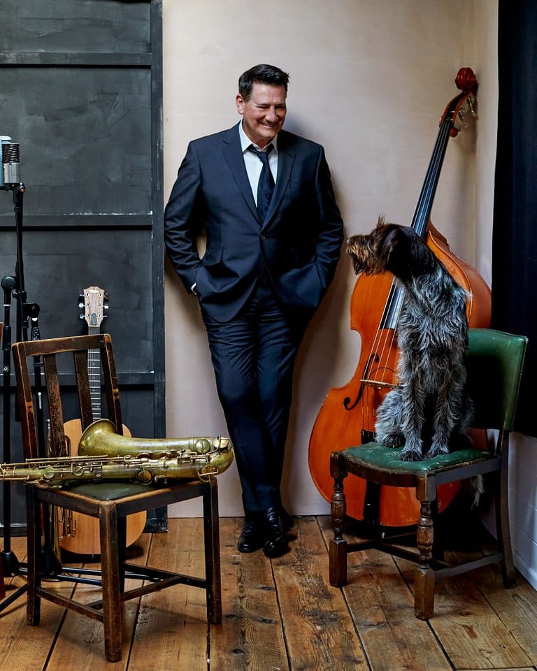 Man in navy suit leaning against wall beside cello, saxophone and microphone in studio setting.