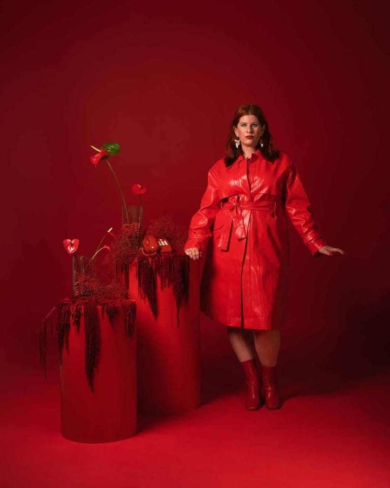 Woman in bright red coat and boots standing beside sculptural floral installation against red background.