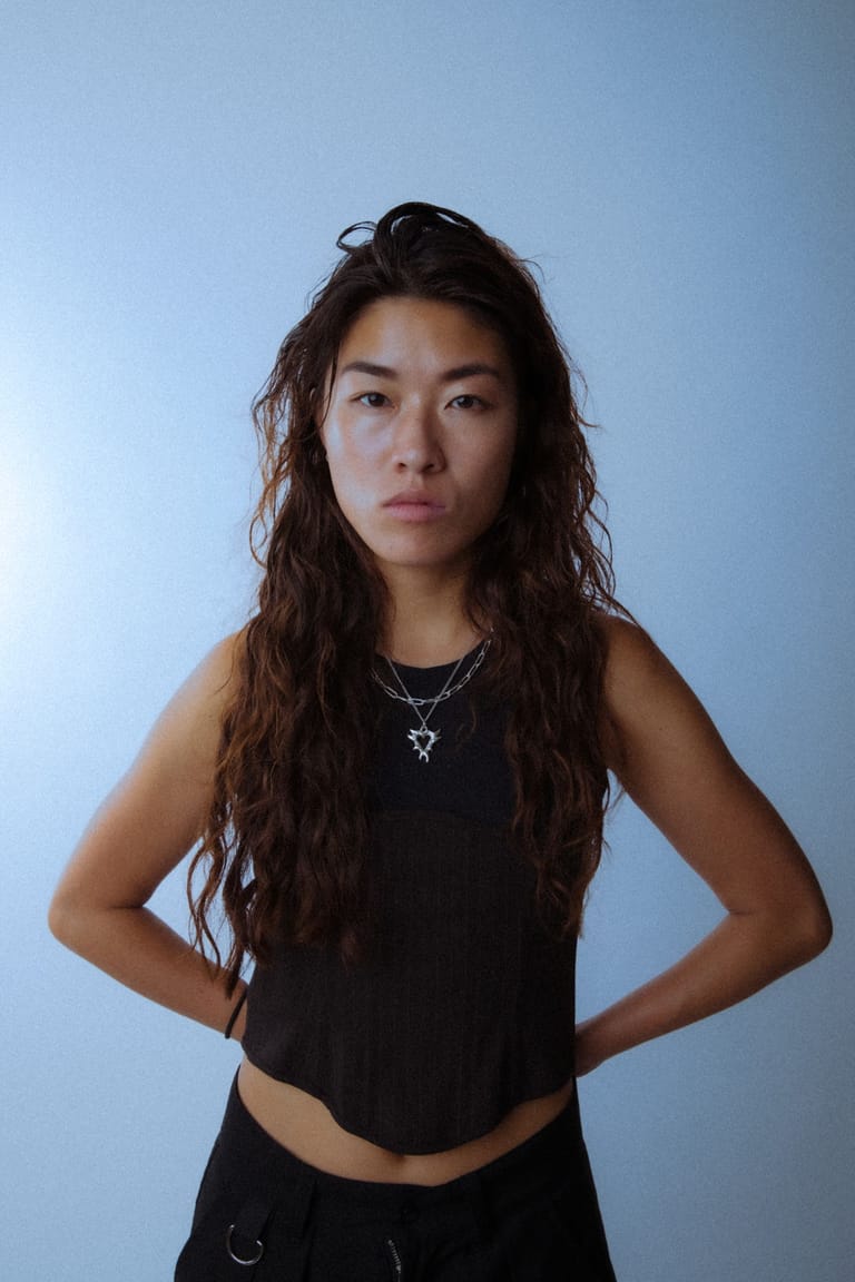 Person with long dark hair wearing black sleeveless top and pendant necklace against blue background.