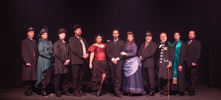 Cast of eleven performers in period costumes standing together on a darkened theatre stage.
