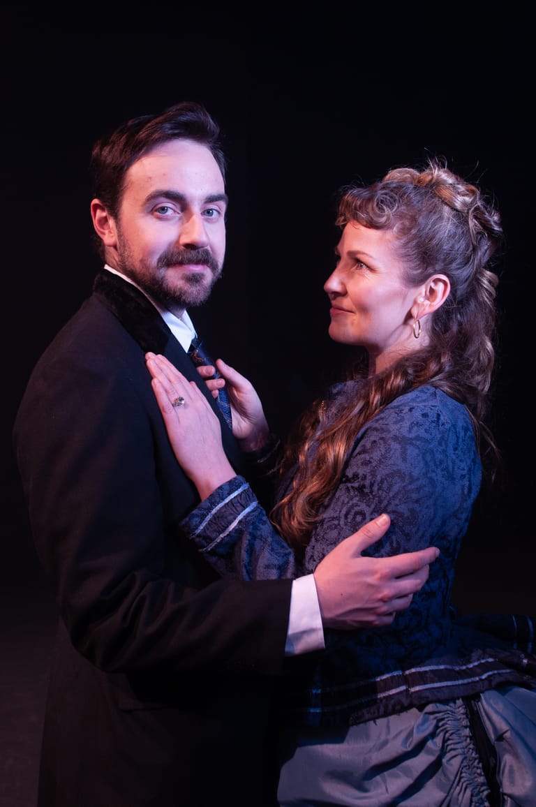 Two performers in period costume embrace on stage against a black background.