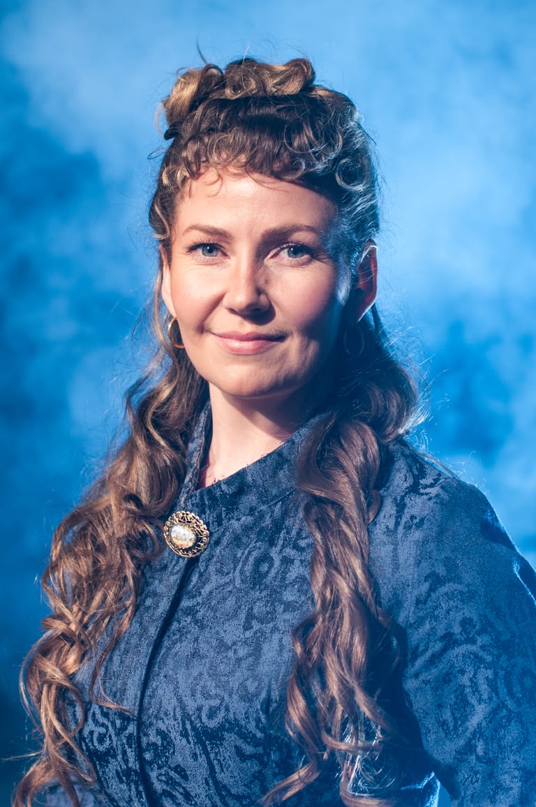 Woman with long braided hair and ornate brooch wearing blue velvet dress against blue backdrop.