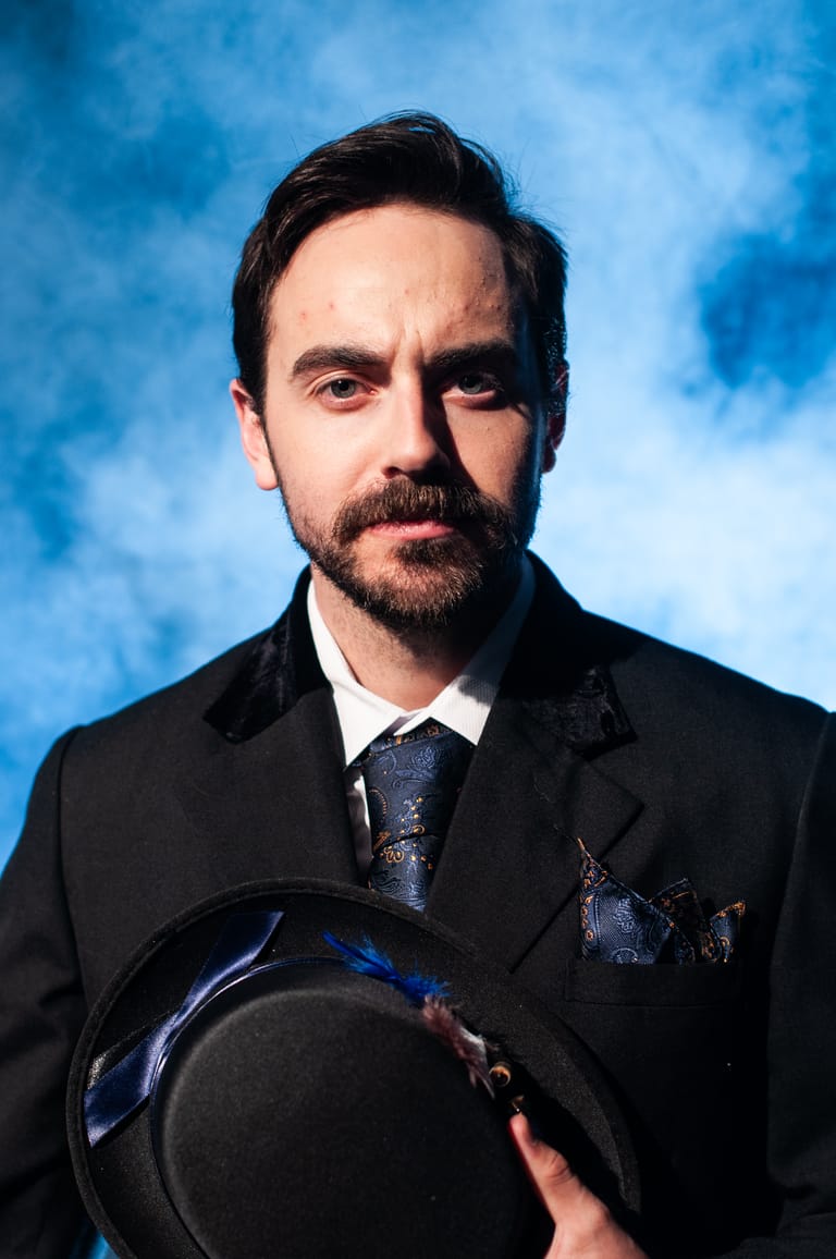 Man in formal black suit and tie holding a dark hat against a blue clouded backdrop.