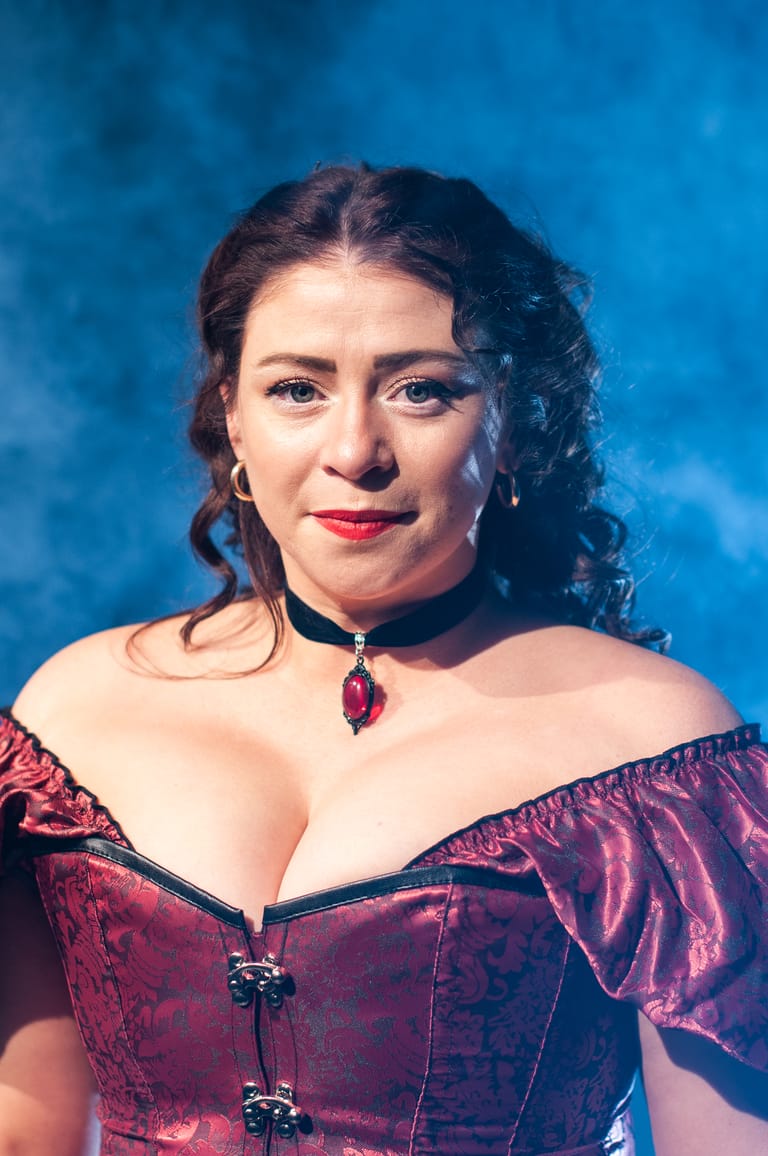 Woman wearing a burgundy off-shoulder corset and jewelled pendant against a blue gradient backdrop.
