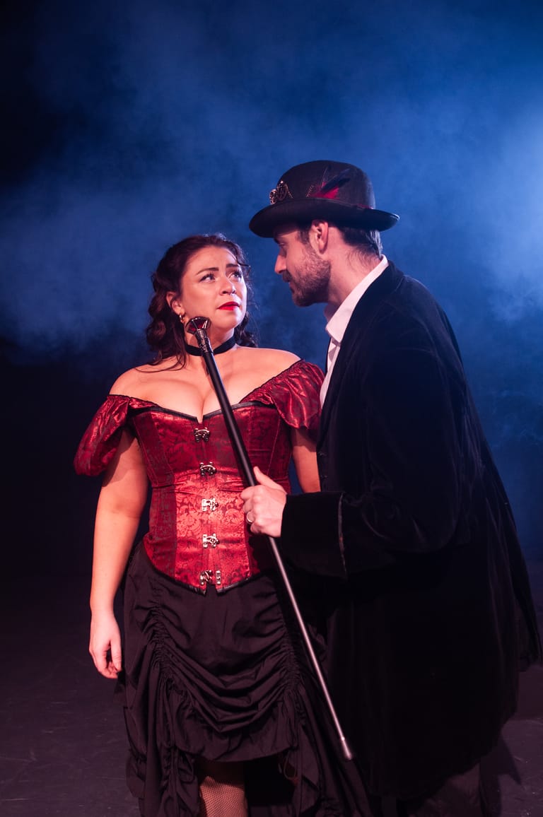 Two performers in Victorian-style costumes stand at a microphone against blue stage lighting, the woman in a red corset looking upward thoughtfully.