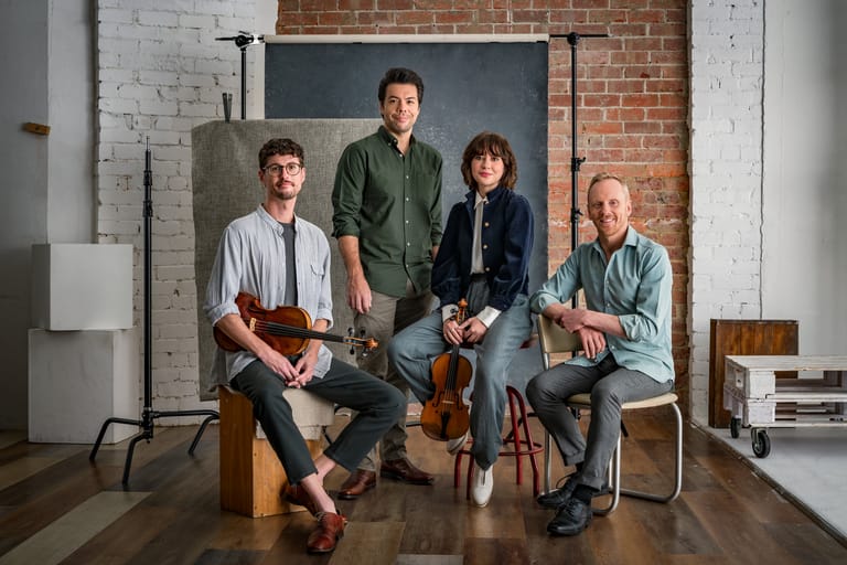 Four musicians holding guitars and bass instruments pose together in a brick studio space.