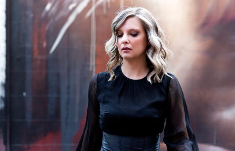 Woman with blonde wavy hair in black clothing against an atmospheric urban backdrop with warm lighting.