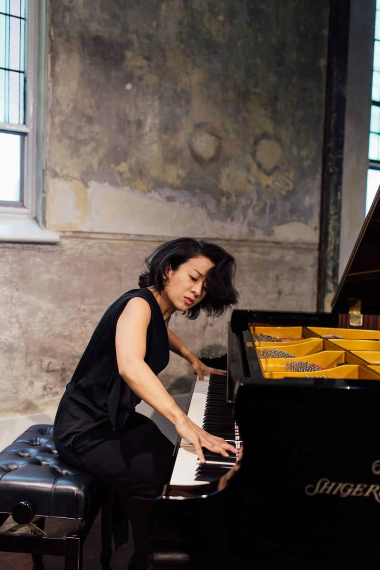 Pianist in black sleeveless dress performing at grand piano in industrial-style venue with weathered concrete walls.