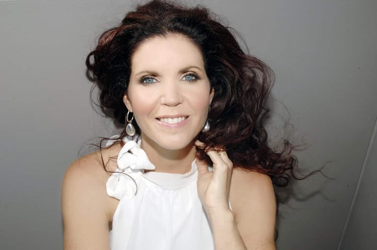 Woman with dark wavy hair wearing a white sleeveless top against a neutral grey background.
