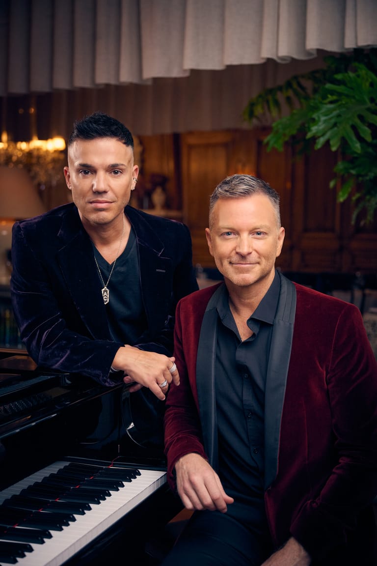 Two men pose beside a grand piano in an elegant interior, one wearing black and the other a burgundy jacket.