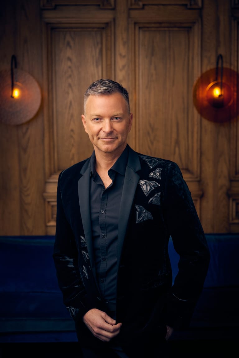 Man in black embroidered jacket stands before wooden doors with warm overhead lighting.