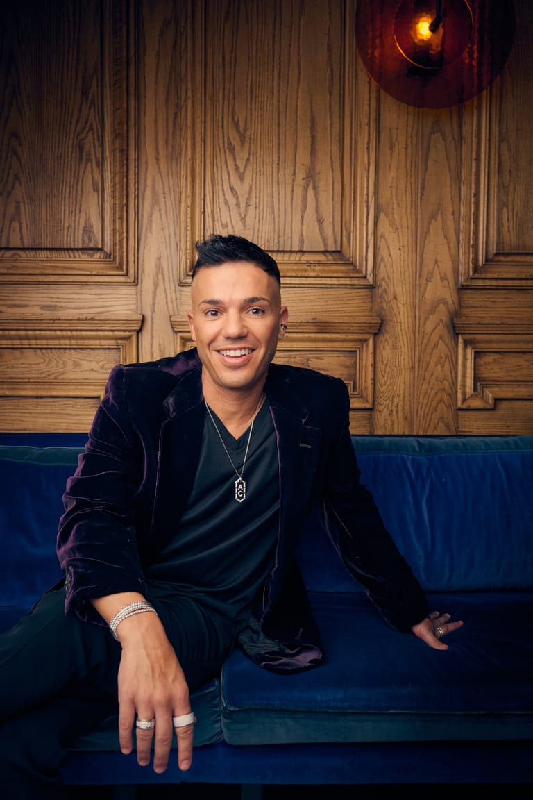 Person in black velvet jacket seated on blue sofa against wooden panelled wall with warm overhead lighting.