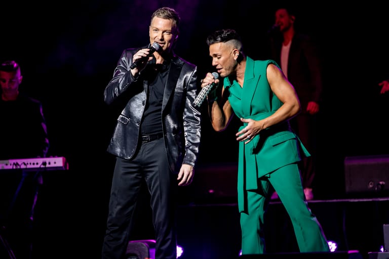 Two performers on stage singing into microphones, one in a dark suit and one in a green sleeveless outfit, bathed in purple stage lighting.