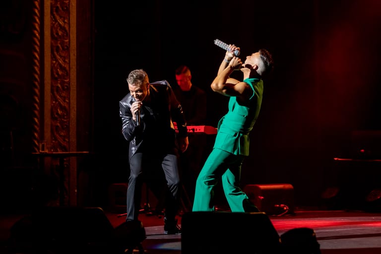 Two performers on stage lit by red spotlights, one singing into a microphone, the other playing a wind instrument in a bright green suit.