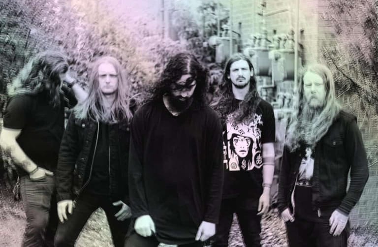 Five band members in dark clothing stand together against a textured, muted-toned backdrop.