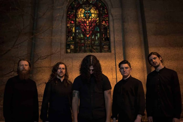 Five musicians in dark clothing stand in a row before an ornate stained-glass window in a stone chapel.