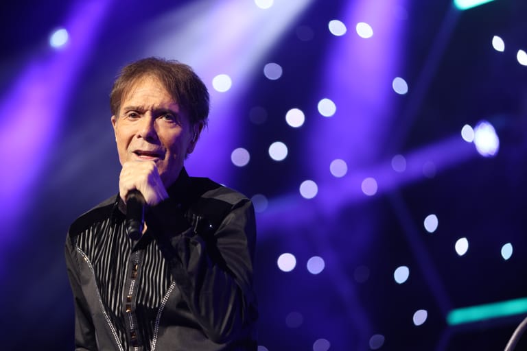 Male performer singing into microphone against purple stage lighting and bokeh background.