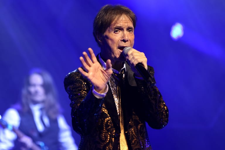 Singer in black and gold jacket performing with microphone under blue stage lighting.