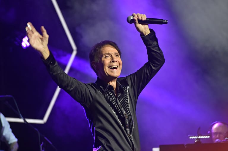 Performer in black jacket singing into microphone with arms raised on purple-lit stage.