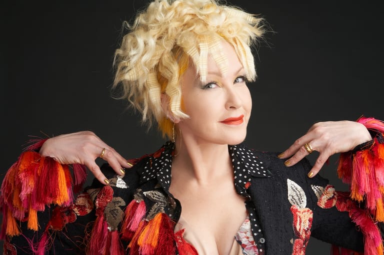 Performer with platinum blonde curled hair wearing a black jacket with vibrant red and orange feathered shoulders against a dark background.
