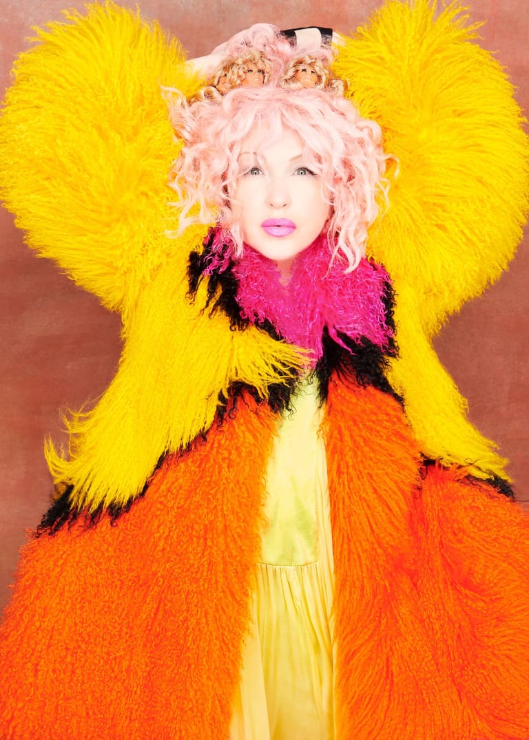 Performer in vibrant yellow, pink and orange fluffy costume with pale pink curled hair against warm-toned backdrop.