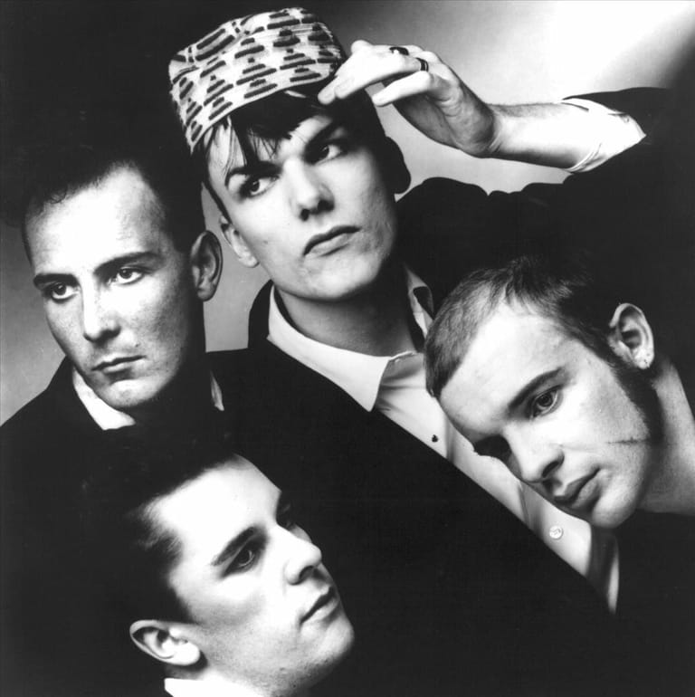 Four men in dark suits pose closely together in black and white portrait, one adjusting a striped headpiece.