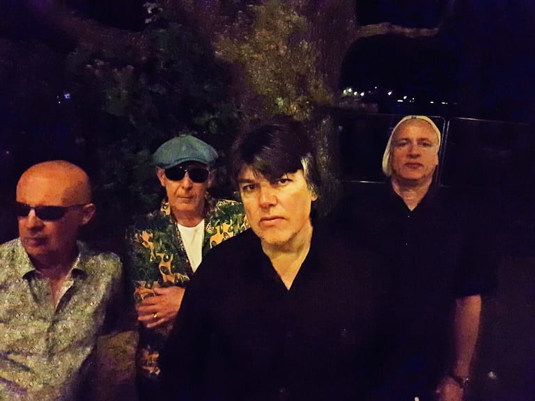 Four musicians posed together outdoors at night, wearing sunglasses and casual attire against a dark, tree-lined backdrop.