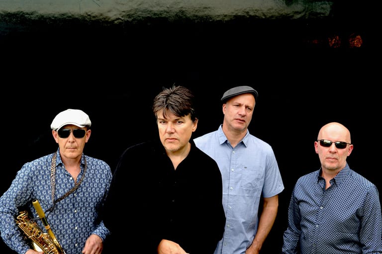 Four musicians posed against a black background, one holding a saxophone.