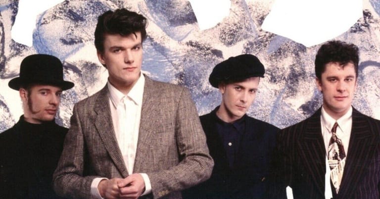 Four men in vintage suits and hats stand against a textured grey backdrop.