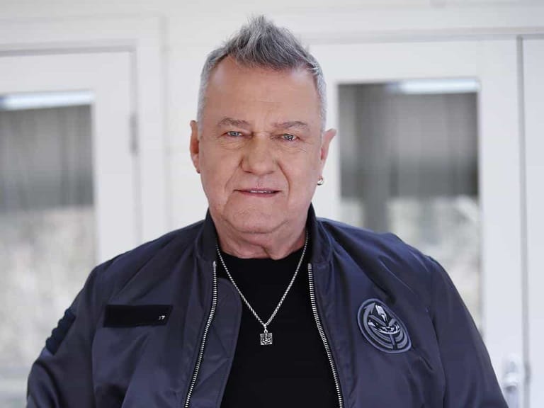 Man with grey spiky hair wearing a dark blue jacket and chain necklace, smiling at the camera indoors.