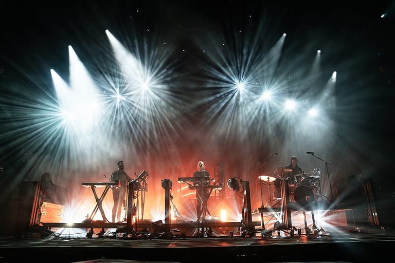 Band silhouetted against dramatic orange stage lighting and radiating white spotlights during live performance.