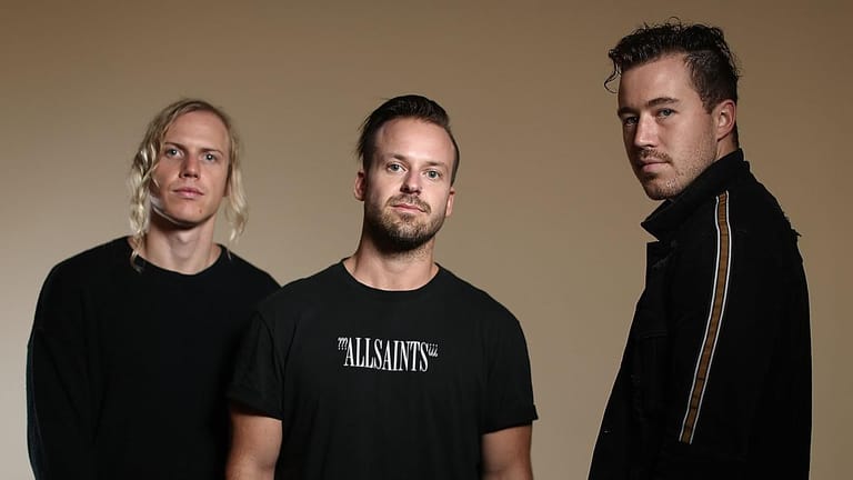 Three men in casual black clothing stand against a neutral beige background in a band promotional portrait.
