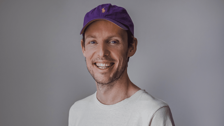 Man in white t-shirt and purple cap smiling at camera against neutral grey background.
