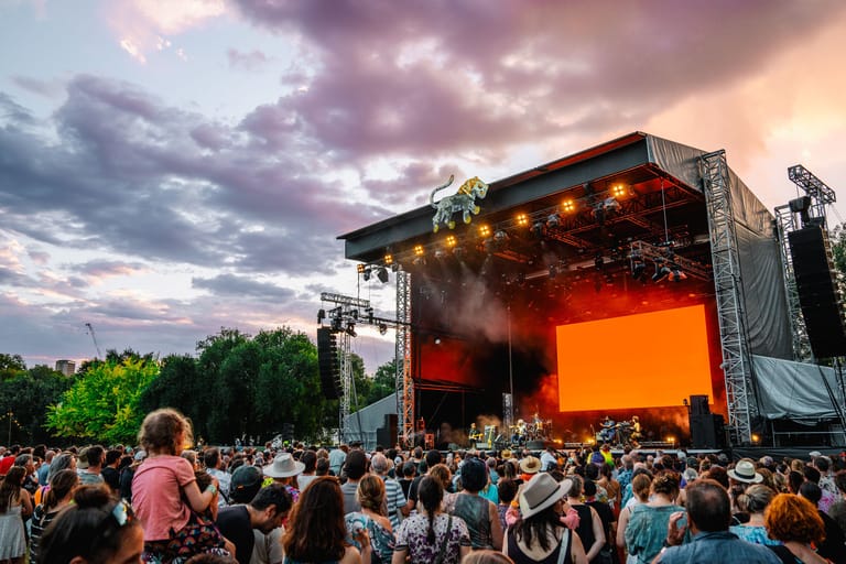 Large crowd gathered before an illuminated outdoor festival stage at dusk beneath dramatic clouds.