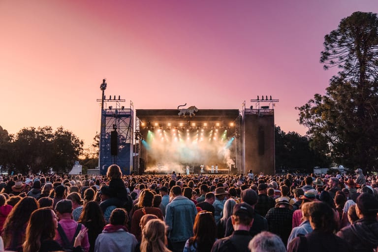 Large crowd watches a stage with bright lights at an outdoor festival during dusk.