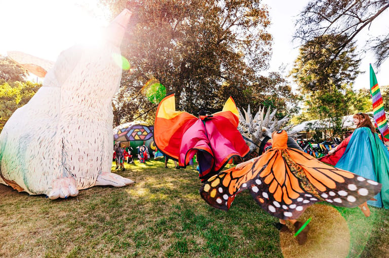 Colourful oversized butterfly and flower sculptures displayed on grass in a sunlit outdoor park setting.