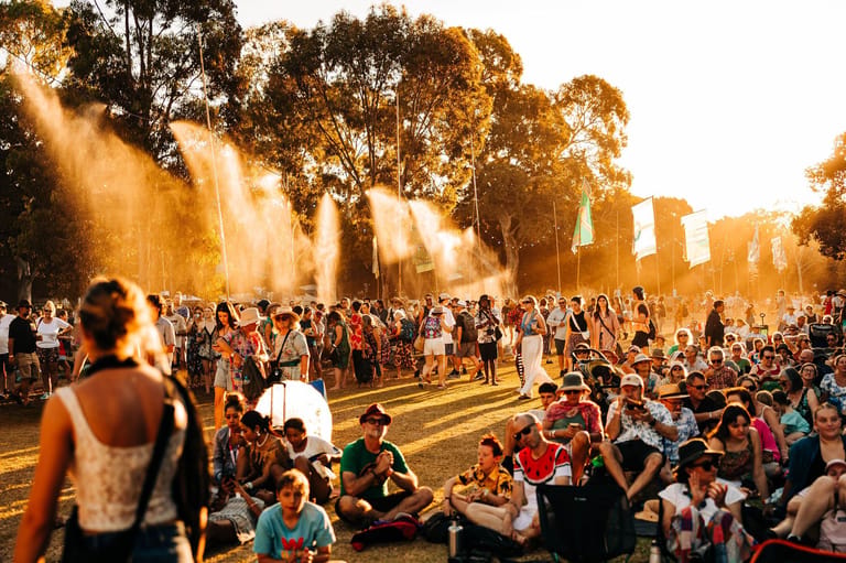 Large crowd gathered outdoors at sunset with water fountains spraying golden light across the park landscape.