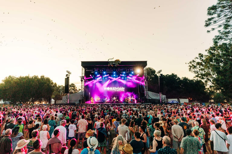 Large outdoor festival crowd facing an illuminated stage with pink and purple lighting at dusk.