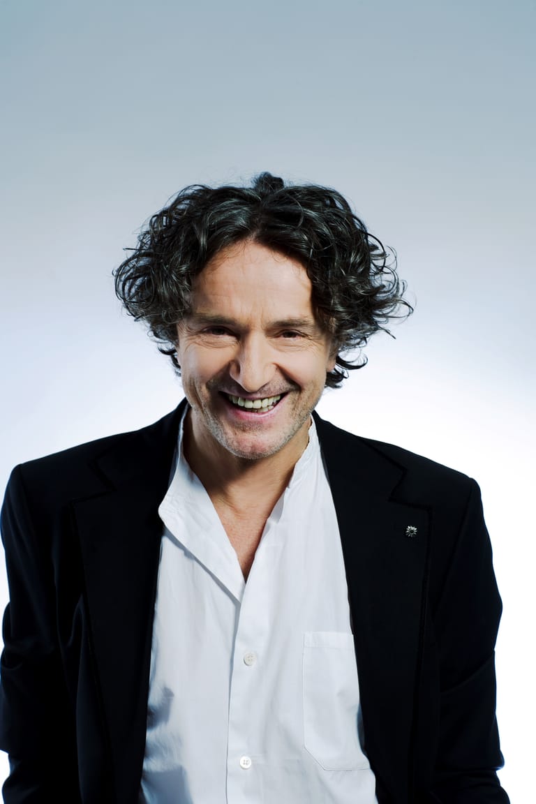 Man with dark curly hair smiling at camera, wearing black blazer and white shirt against light blue background.