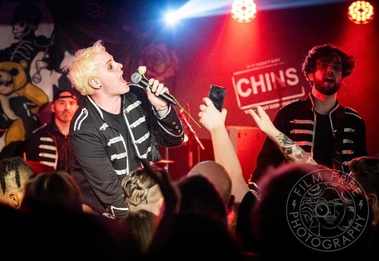 Singer in skeleton-patterned black outfit performing at microphone on stage with red lighting and band members visible.