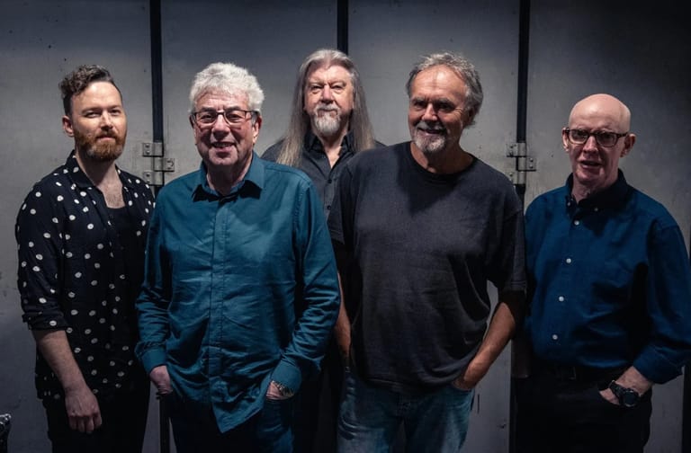 Five men standing together against a grey wall, dressed in casual shirts and sweaters.