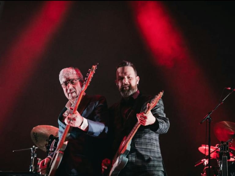 Two guitarists perform on stage beneath red spotlights during a live concert.
