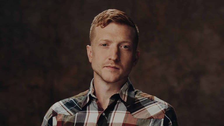 Man in a plaid shirt against a dark textured background, looking directly at camera.