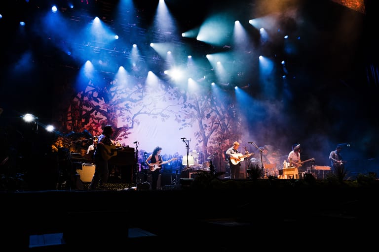 Band performing on stage with blue and orange lighting, bare branches set piece, and audience silhouettes in foreground.