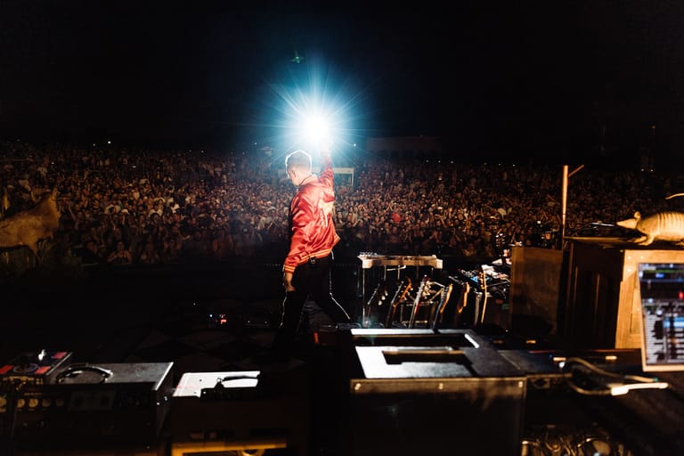 Performer in red jacket on stage before a large crowd illuminated by bright overhead lighting at night.